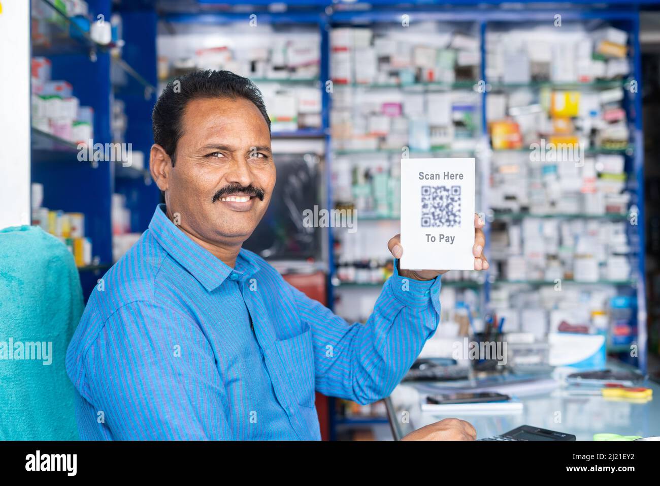 Sorridente medico proprietario mostrando scansione qui per pagare qr code board per il pagamento guardando la macchina fotografica alla farmacia - concetto di pagamento online, wireless e. Foto Stock