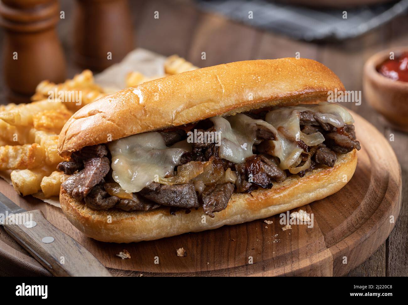 Sandwich di cheesesteak di Philly preparato con bistecche, formaggio e cipolle su un rotolo di hoagie tostato con patatine fritte su un asse di legno Foto Stock