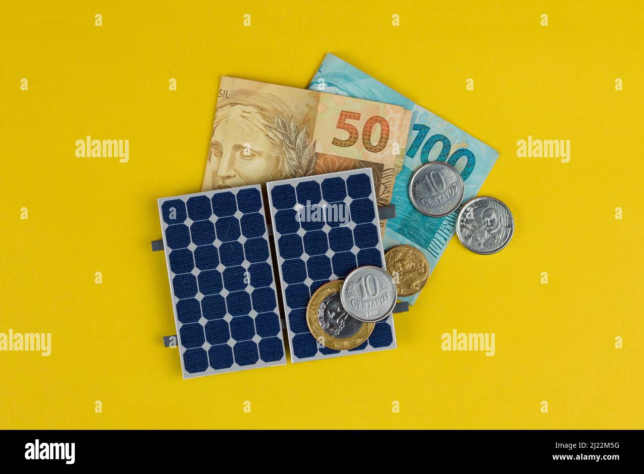 Pannello fotovoltaico, denaro e filo di spina isolati su sfondo giallo. Pannello olar con vari valori di denaro brasiliano. Bus energia pulita alternativo Foto Stock