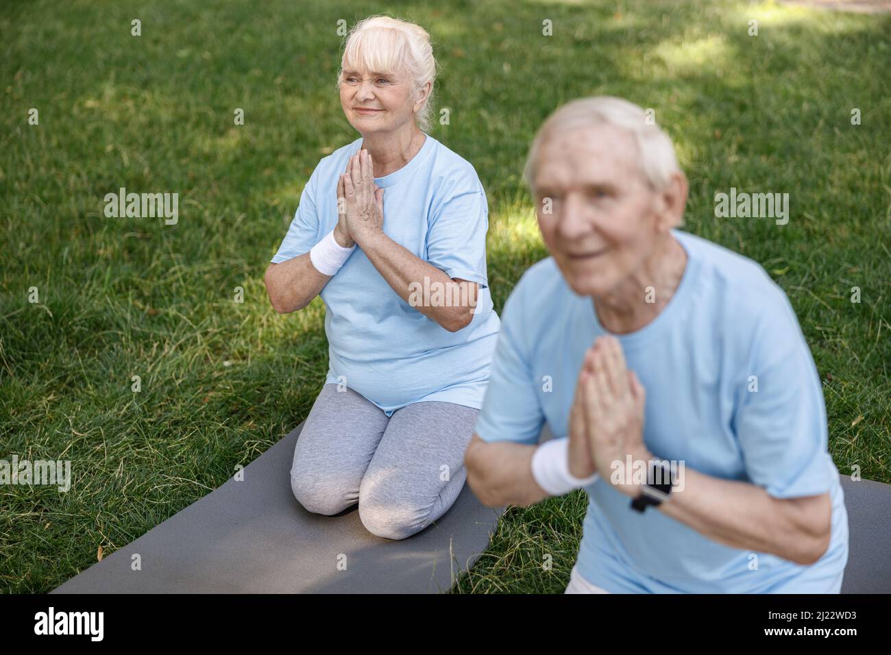 Sorridente donna anziana con marito fare namaste praticare yoga sul prato in parco Foto Stock
