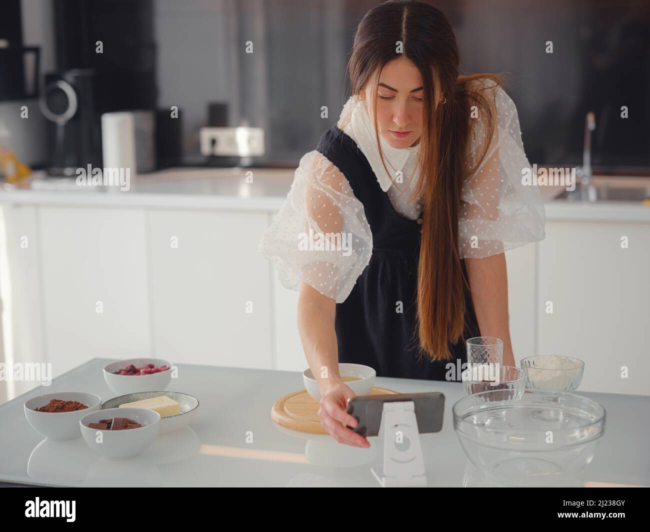 Giovane donna guarda una ricetta per cucinare una torta in uno smartphone. Tutorial di cibo o corsi di cucina online. Dolce concetto di cottura. Foto Stock