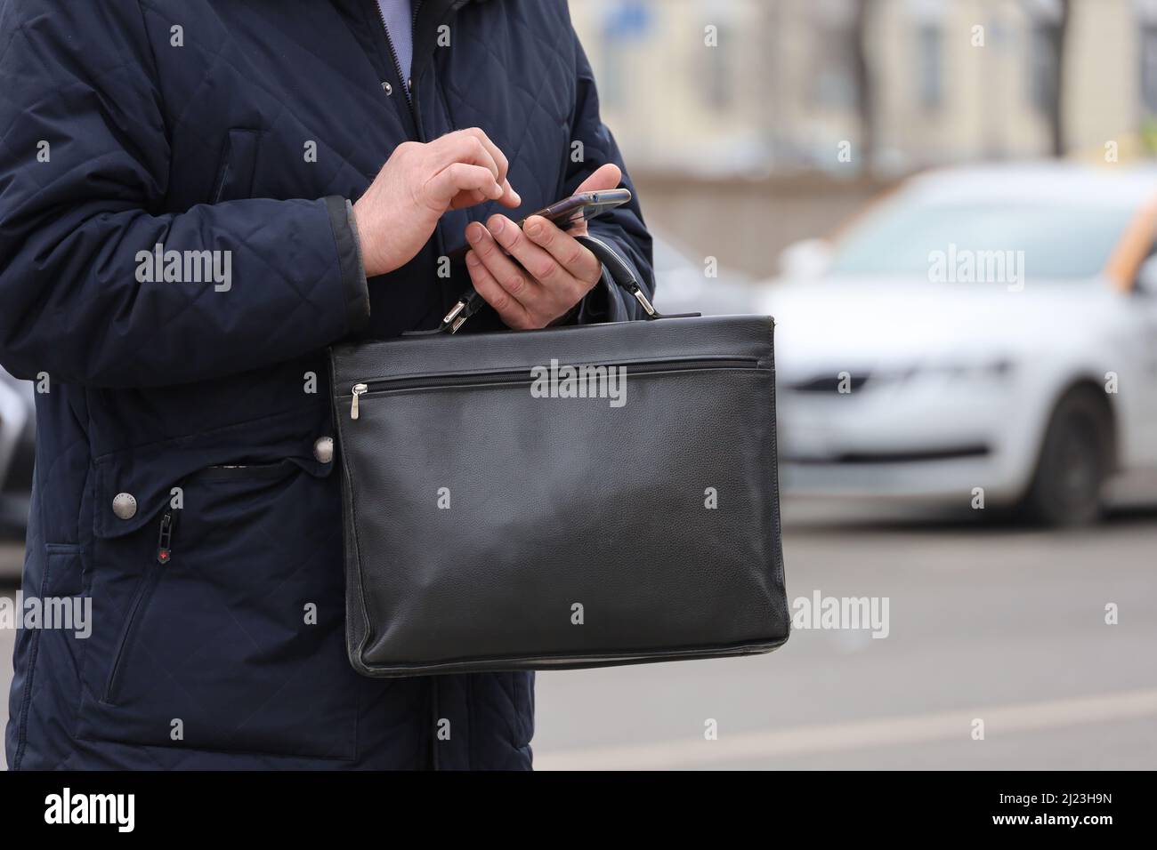 Uomo in cappotto in piedi con una borsa portadocumenti in pelle con smartphone in mano su una strada cittadina. Concetto di uomini d'affari, funzionari, dipendenti governativi Foto Stock
