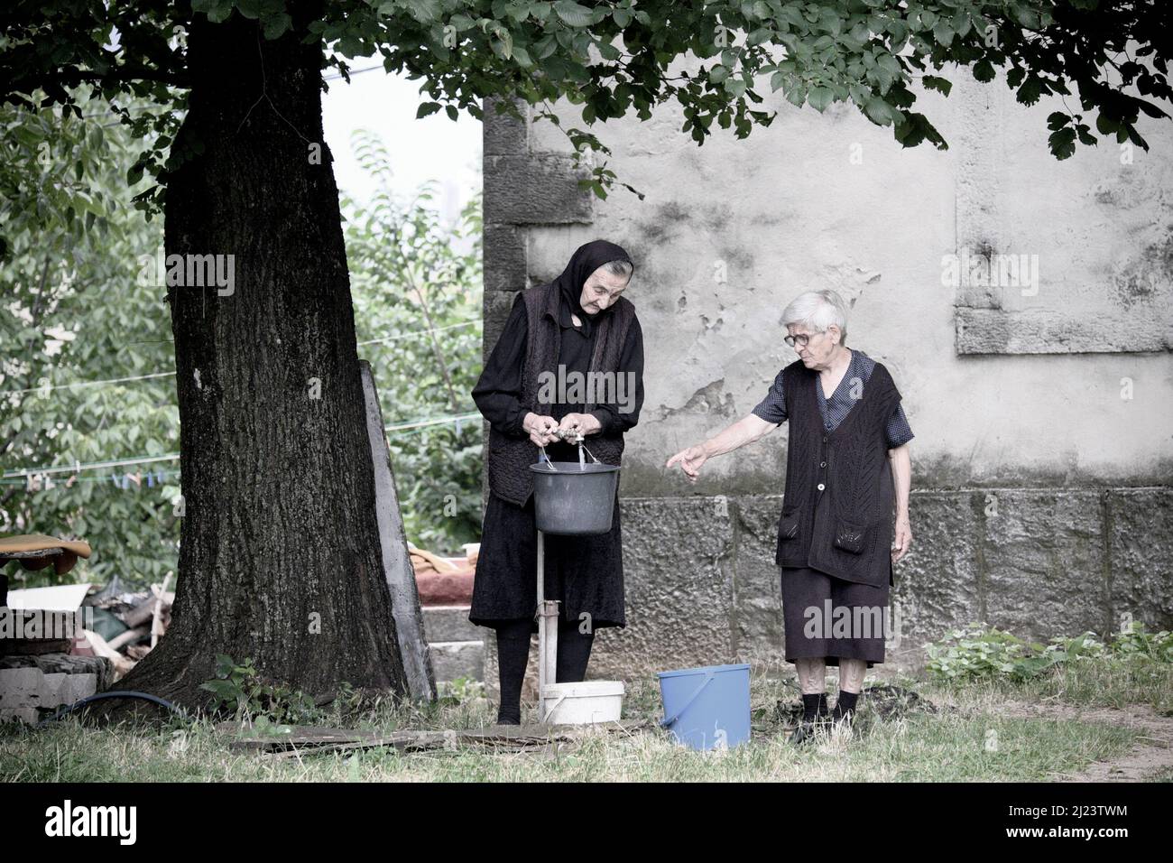 NIKSIC, MONTENEGRO - 25 LUGLIO 2016: Due donne anziane in abiti neri stanno riempiendo alcuni secchi con l'acqua di un rubinetto all'aperto sotto un grande albero Foto Stock
