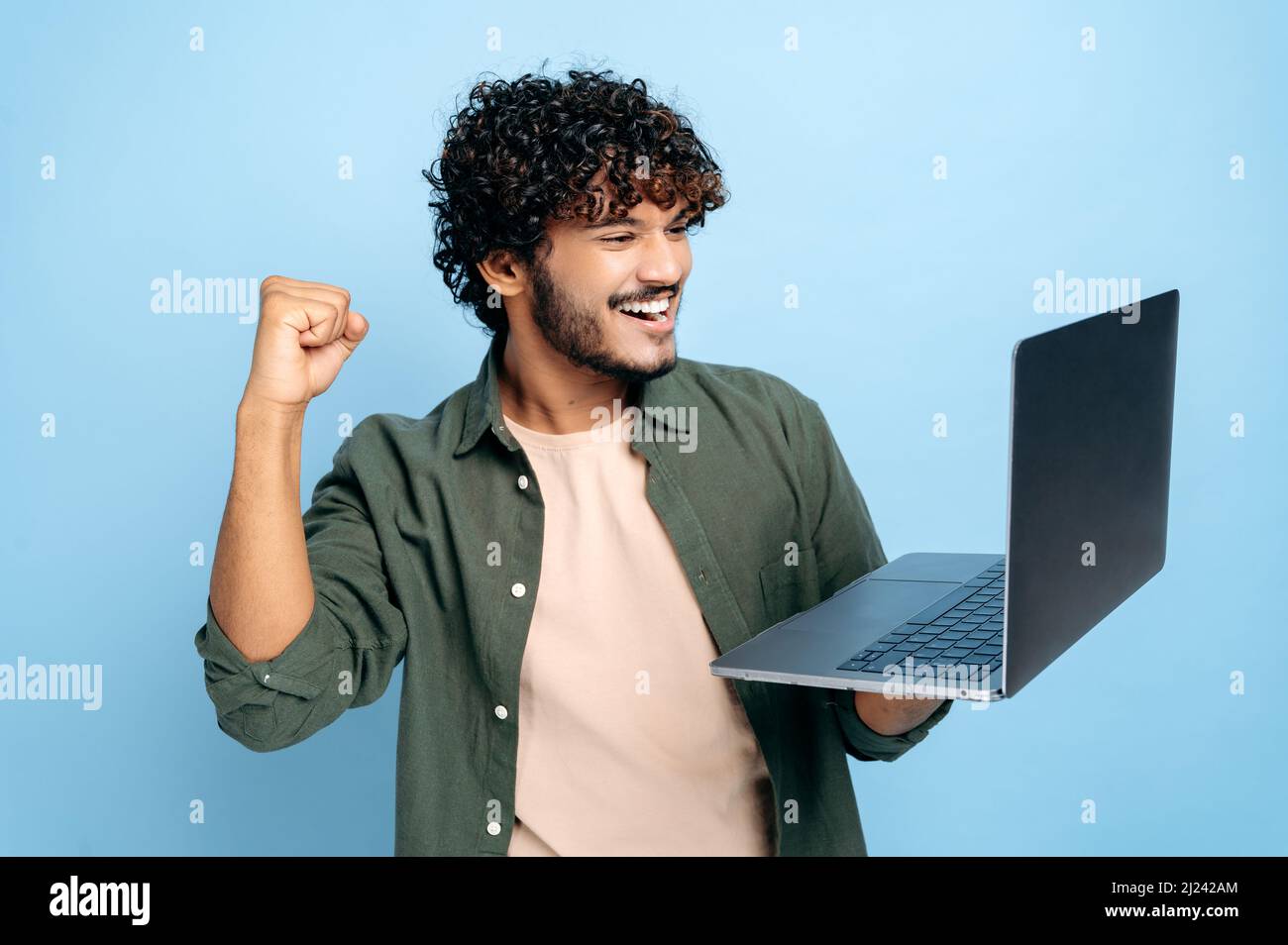 Gioioso ragazzo arabo felice o indiano in abiti casual, studente o freelancer, tiene un laptop aperto in mano, si rallegra nella vittoria, gesturing con il suo pugno, in piedi su un isolato sfondo blu Foto Stock
