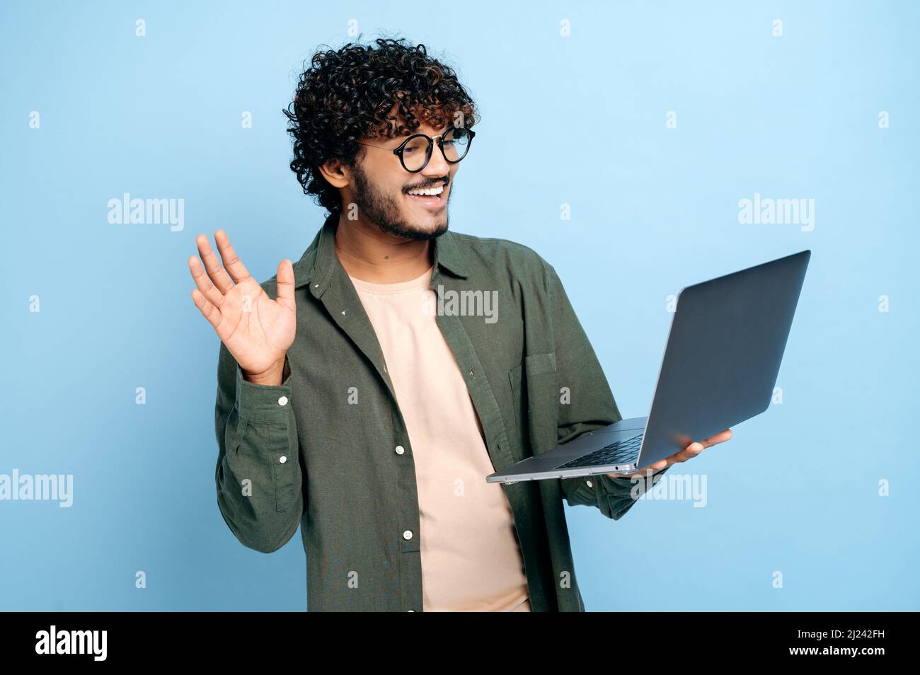 Comunicazione video online. Il tipo indiano o arabo, in abiti casual, freelancer o studente, utilizza un computer portatile per una videoconferenza, si erge su sfondo blu isolato, saluta un interlocutore, sorridendo Foto Stock