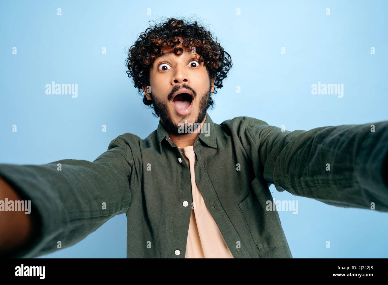 Divertente ragazzo indiano positivo gioioso con gladden volto espressione, in camicia casual, facendo selfie girato su smartphone e fotografando intorno nella fotocamera, si trova su isolato sfondo blu Foto Stock