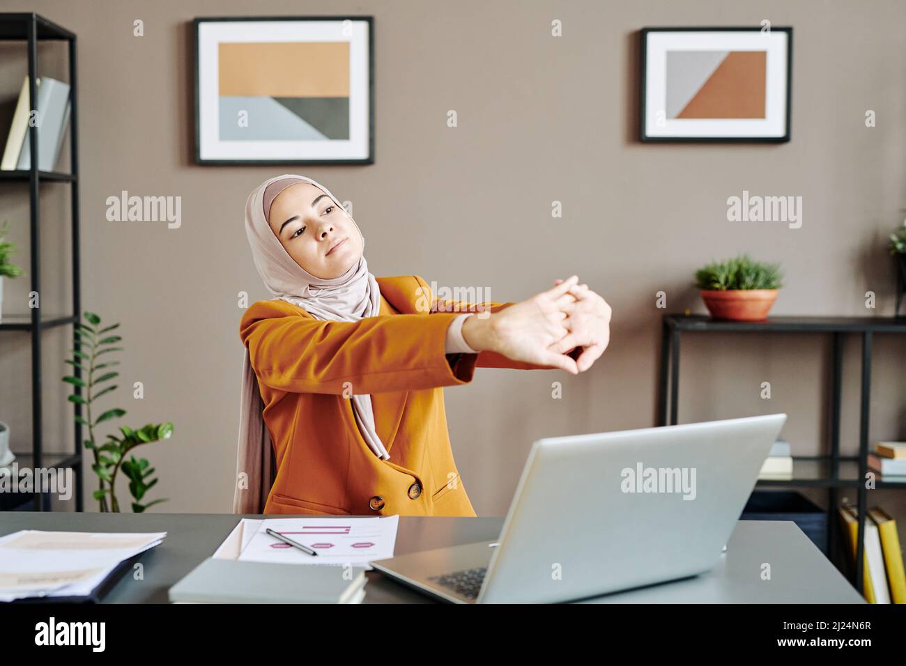 Giovane lavoratrice in ufficio stanca che tiene le braccia tese di fronte a se stessa mentre fa l'esercizio davanti al computer portatile dalla scrivania Foto Stock