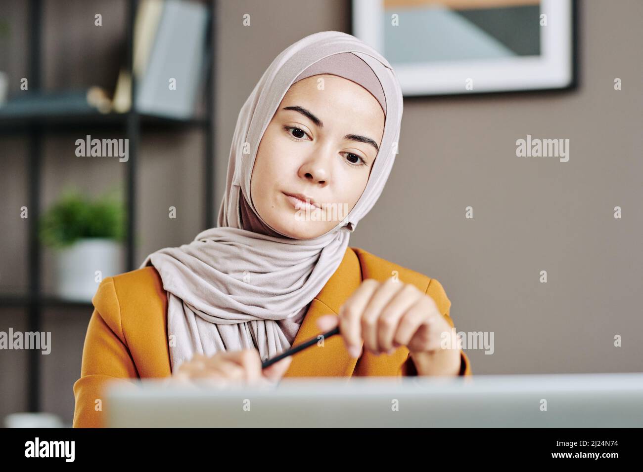 Giovane donna musulmana seria in hijab seduta davanti al portatile in ufficio e guardando il partner commerciale sullo schermo durante la riunione digitale Foto Stock