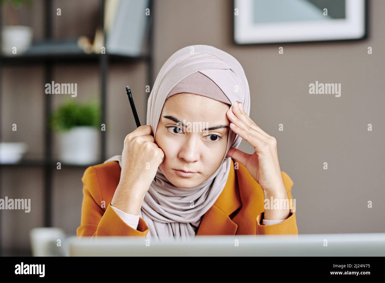 Una giovane e seria dipendente in hijab che guarda attentamente lo schermo del laptop durante un incontro digitale con il partner aziendale Foto Stock