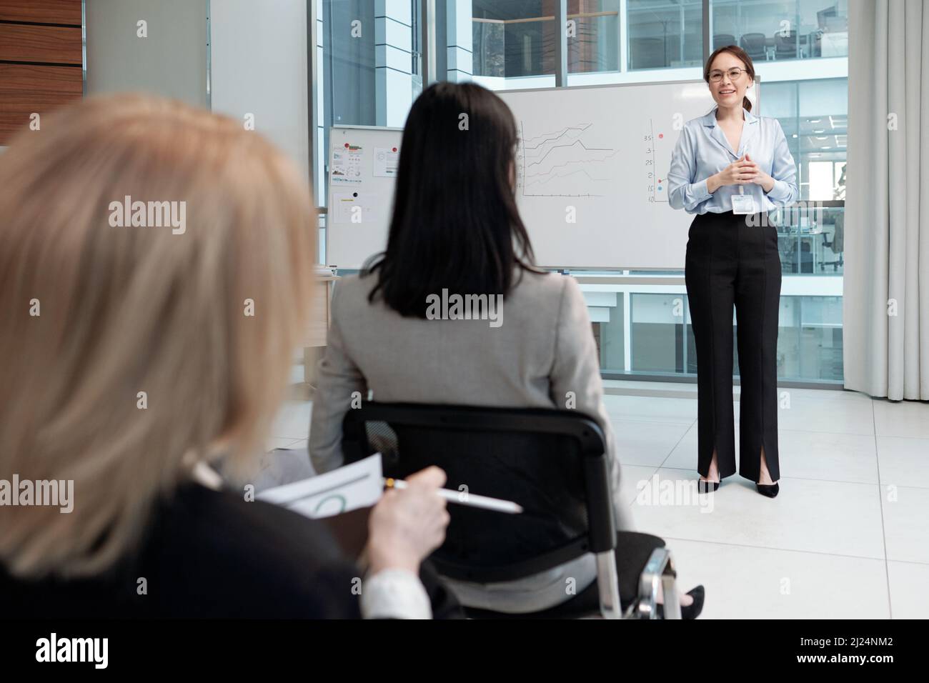 Giovane vettura sorridente di affari in formalwear che comunica con il pubblico al seminario mentre si levano in piedi dalla lavagna bianca con i grafici Foto Stock