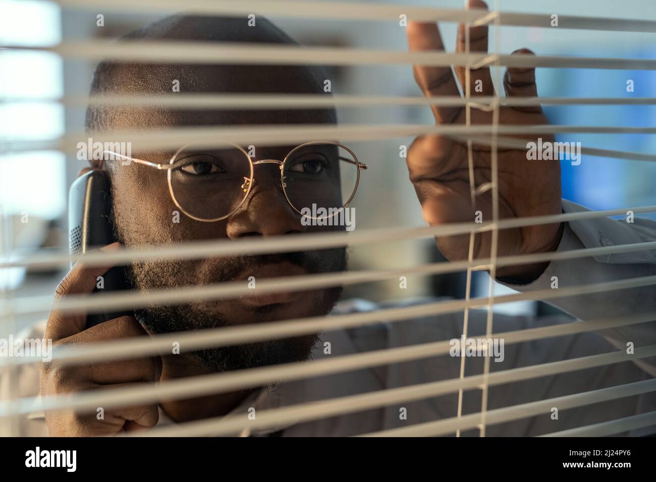 Faccia dell'ufficiale afro-americano negli occhiali che parla sul telefono cellulare mentre osserva attraverso le persiane veneziane nel reparto di sicurezza Foto Stock