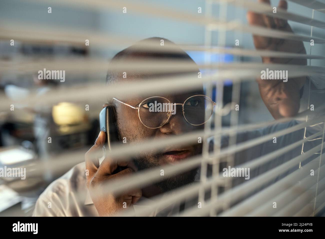 Il personale del dipartimento del sistema di sicurezza maschile afro-americano parla al telefono mentre guarda attraverso le tende veneziane in ufficio Foto Stock