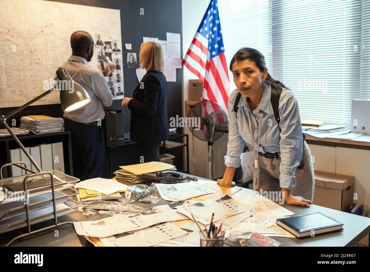 Giovane donna ispanica grave detective piegarsi sul posto di lavoro con documenti sullo sfondo di due colleghi di comunicazione Foto Stock