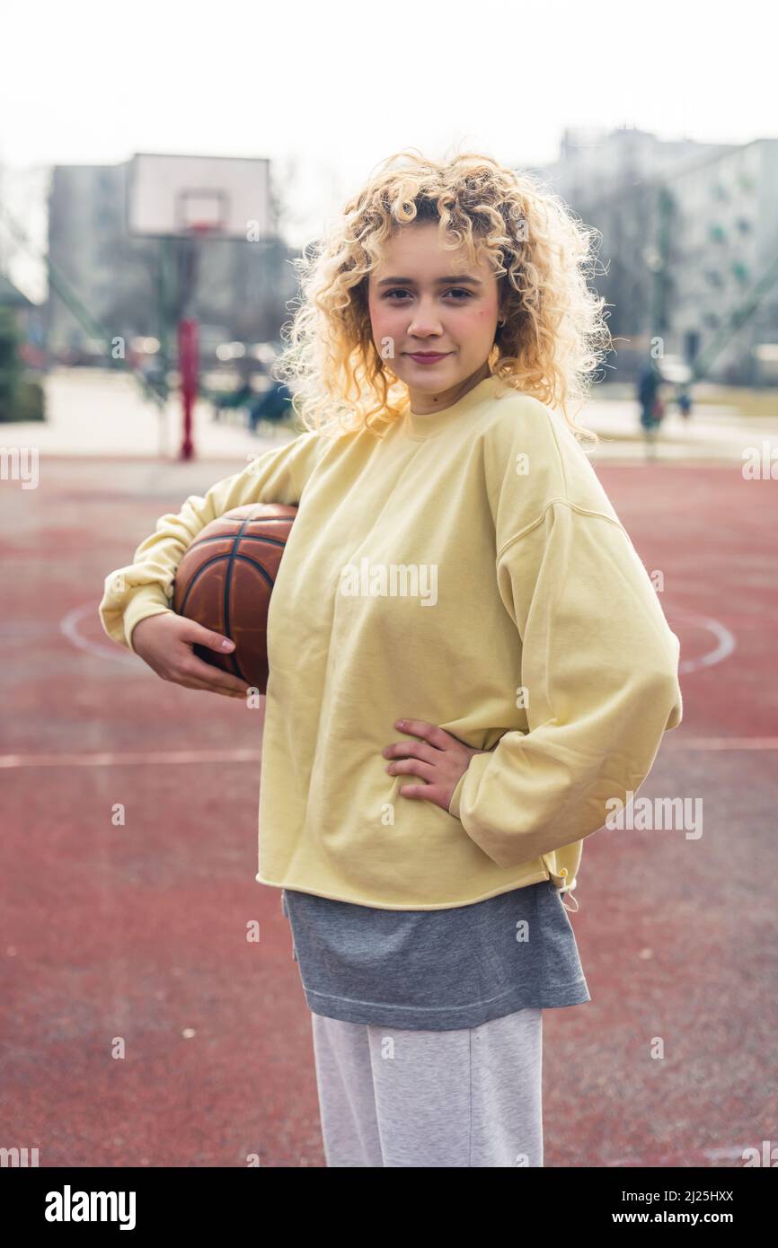 Sicura giovane bionda caucasica donna si erge in un maglione giallo chiaro sul campo sportivo tenendo una palla di basket e mantenendo un'altra mano sul suo girovita medio pieno colpo . Foto di alta qualità Foto Stock