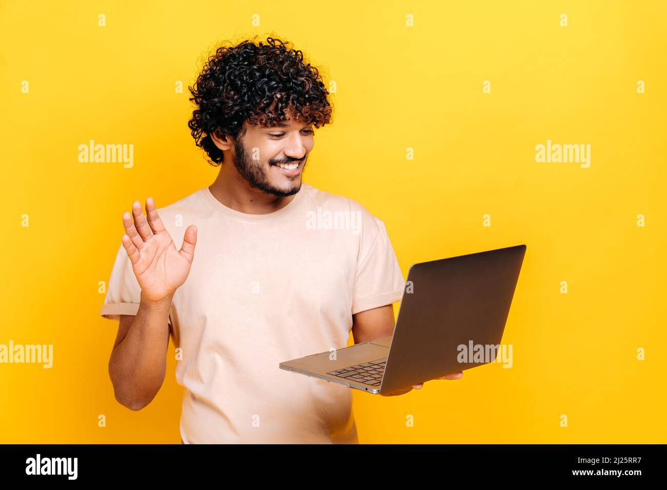 Positivo giovane adulto indiano o arabo ragazzo tenere un laptop, parlare in videoconferenza online, saluto interlocutore, sorridente amichevole, in piedi su sfondo isolato arancione in T-shirt casual Foto Stock