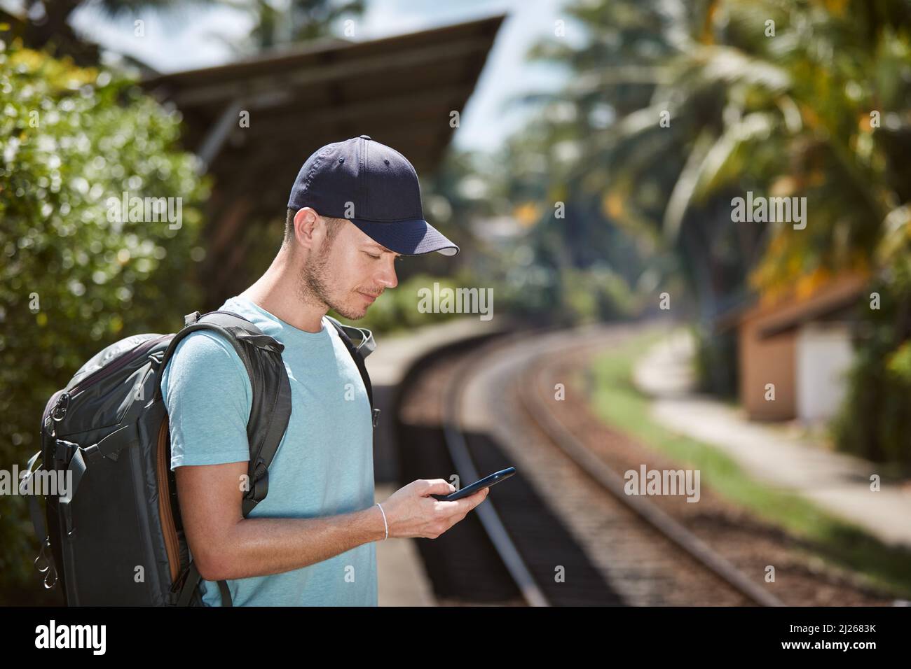 Turista con zaino in destinazione tropicale di viaggio. Vista laterale dell'uomo che usa il telefono cellulare in attesa del treno alla stazione ferroviaria. Foto Stock