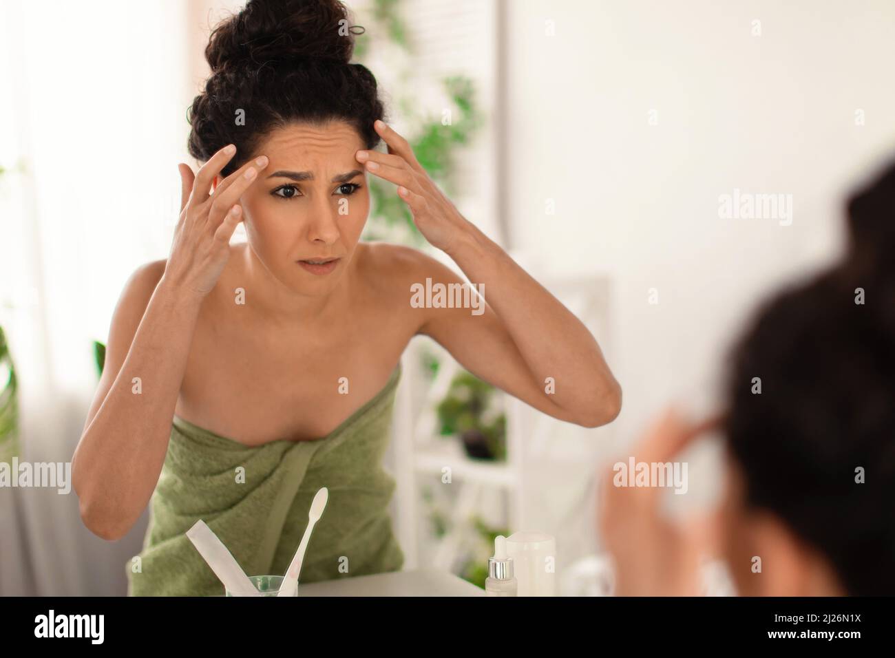 Preoccupata donna millenaria guardando la sua riflessione nello specchio, controllando le prime rughe sulla fronte in bagno, spazio libero Foto Stock