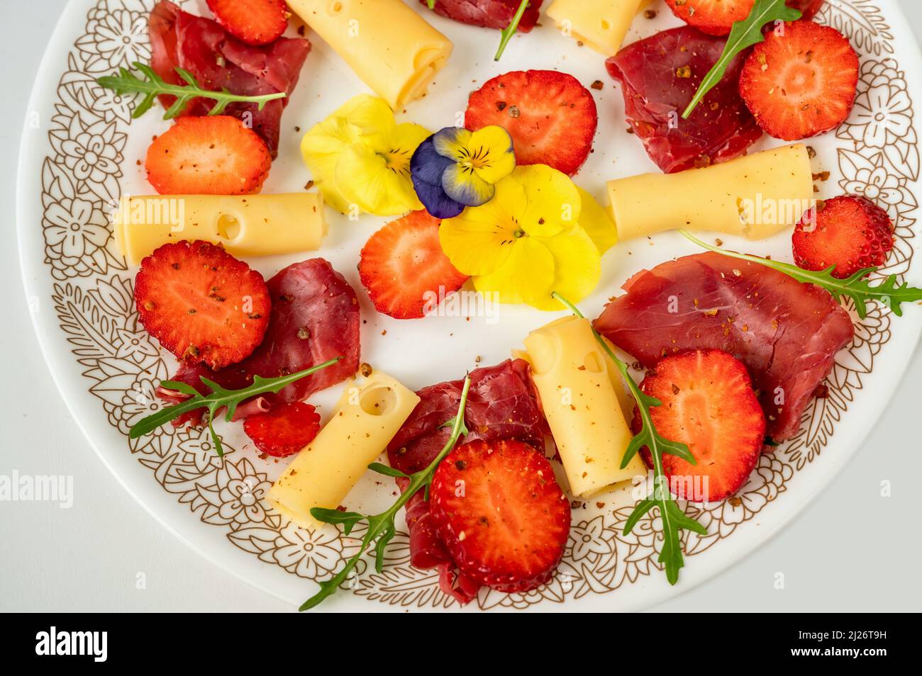 Antipasto leggero con fragola, formaggio e carpaccio, testa di fiori di pancia commestibile e foglia di rucola sul piatto, primo piano. Foto Stock