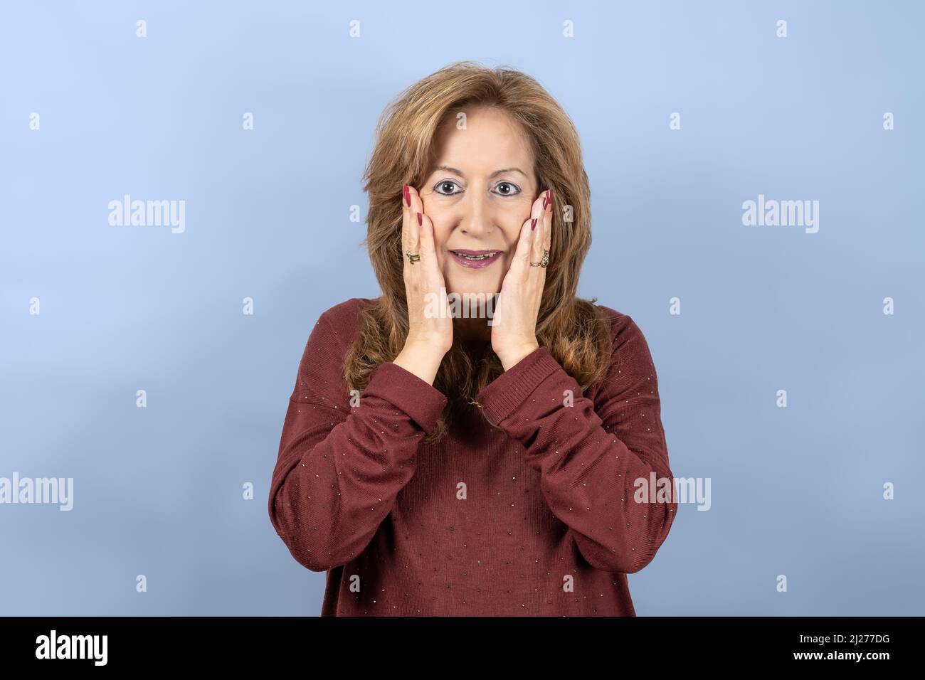 Donna con un volto sorpreso e le mani sul suo volto per l'emozione, sfondo blu studio. Foto Stock