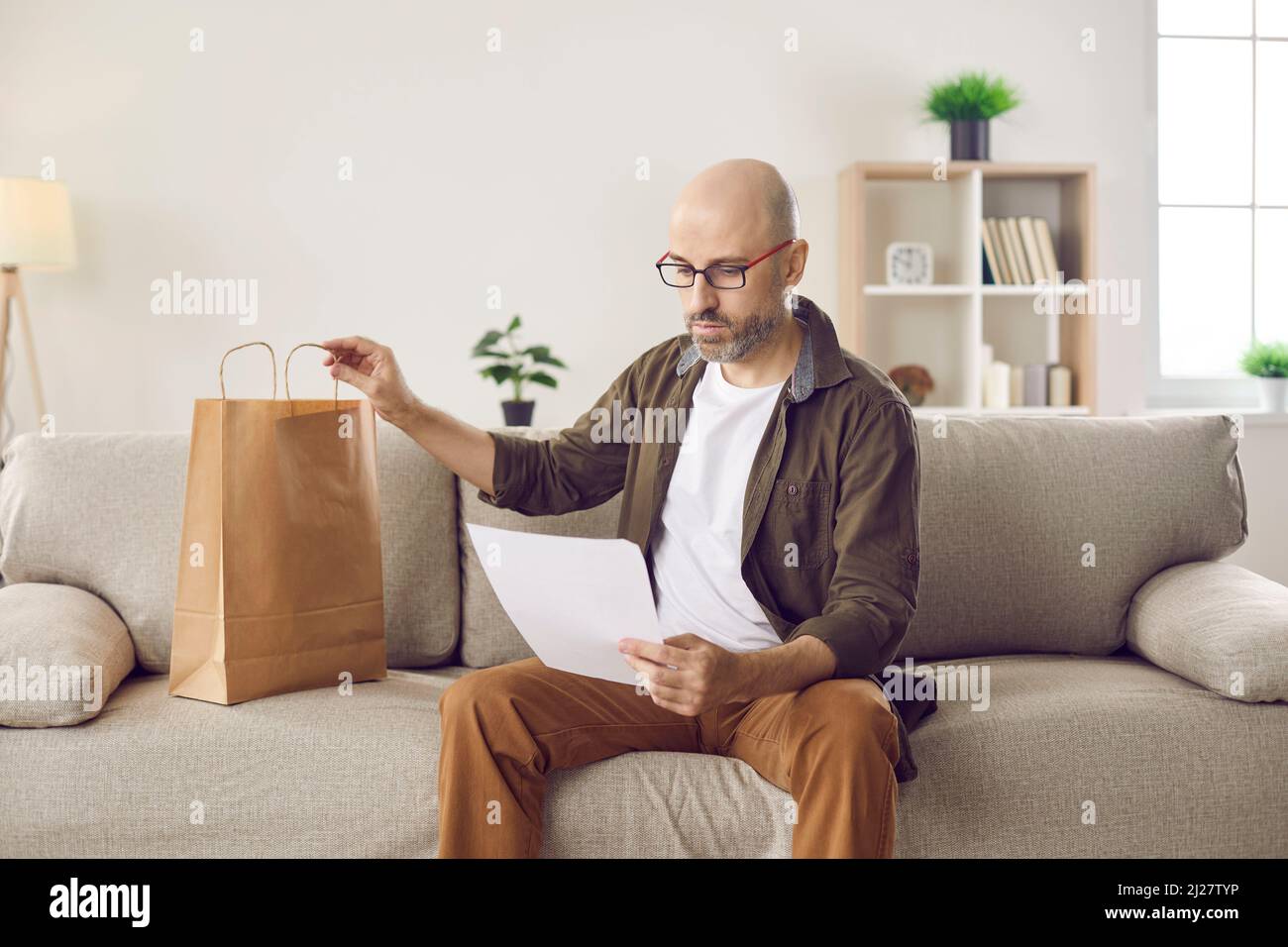 Uomo seduto sul divano a casa e la lettera di lettura si è Unito con la borsa di consegna di carta Foto Stock