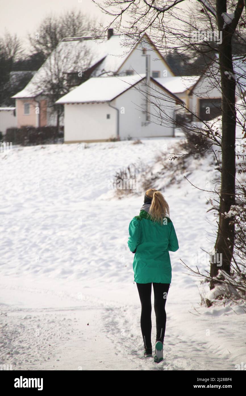 Una vista posteriore verticale di una runner femminile in una giacca verde in un parco innevato Foto Stock