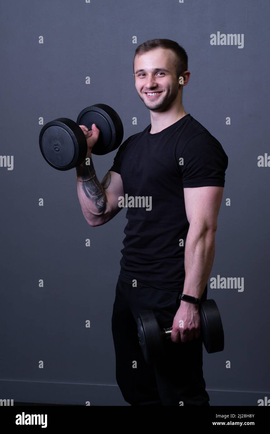 Un ragazzo con manubri in una T-shirt nera muscolare uomo sport in posa, da grigio invecchiato in ragazzo e allenamento atletico, caucasico attraente. Attivo Foto Stock