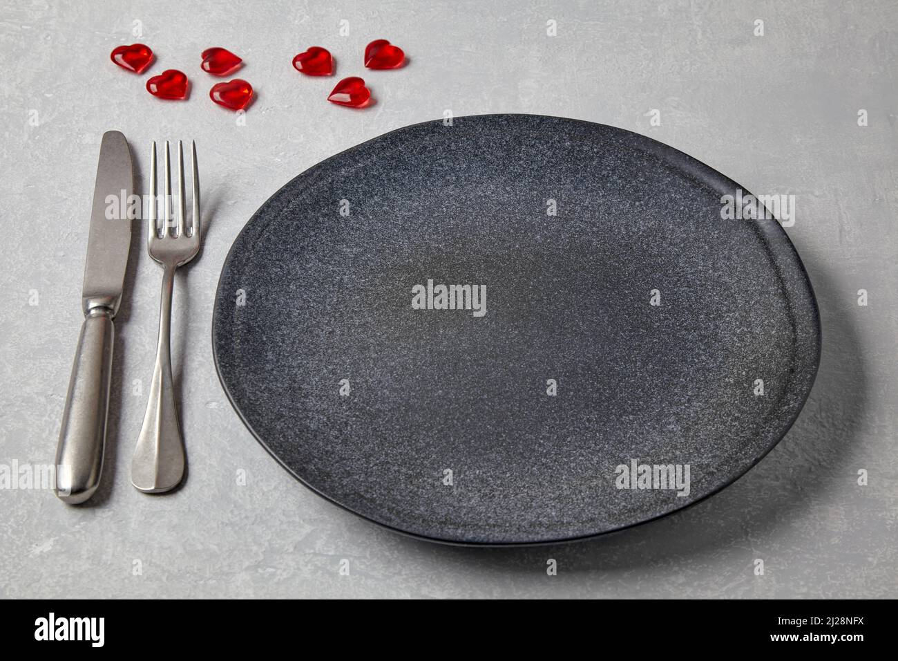 Piatto scuro vuoto con posate e cuori di caramello rosso su un tavolo di cemento chiaro Foto Stock