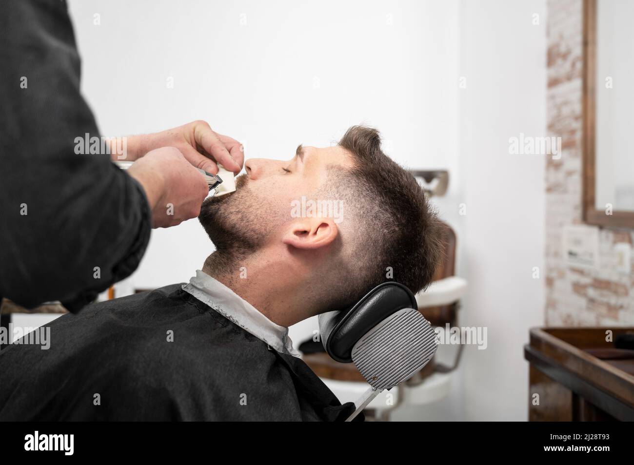 Giovane ippico uomo caucasico durante la cura della barba in un moderno barbiere. Styling per capelli da uomo. Bell'uomo che ottiene un nuovo acconciatura con rifinitore elettrico. Fotografia di alta qualità Foto Stock