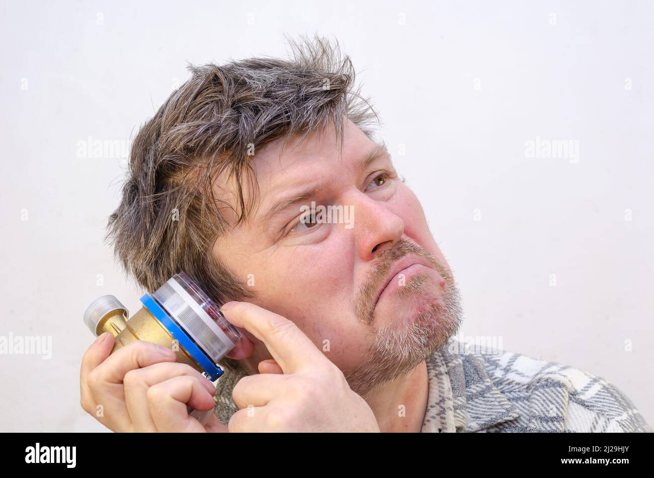 Il maschio ascolta il contatore dell'acqua con l'orecchio. Uomo con capelli aggrovigliati e diseveleati davanti ad una parete di colore grigio chiaro. All'interno. Messa a fuoco selettiva. Foto Stock