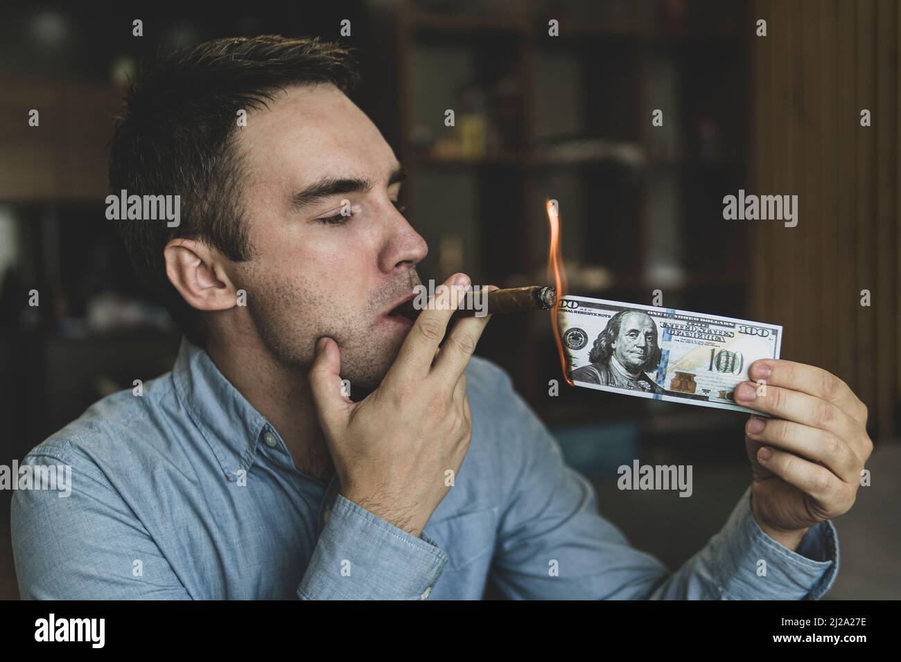 Giovane brutale uomo d'affari che illumina sigaro con 100 dollari di fattura come simbolo di ricchezza e successo. Il concetto di ricchezza e stravaganza. Foto Stock