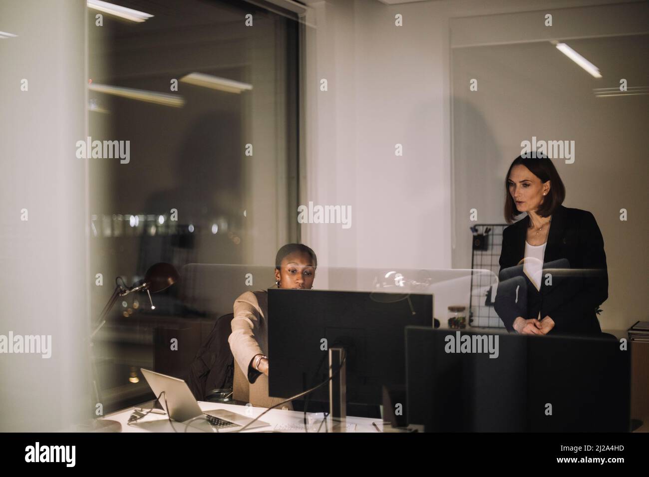 Collega di sesso femminile che parla con una donna d'affari sul computer visto attraverso un bicchiere d'ufficio Foto Stock