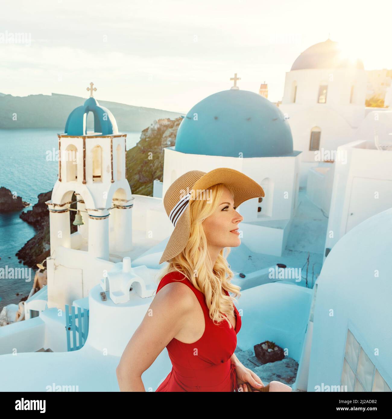 Donna bionda che indossa un abito rosso estivo e un cappello da sole, con le famose cupole blu della chiesa sullo sfondo, a Oia a Santorini Foto Stock