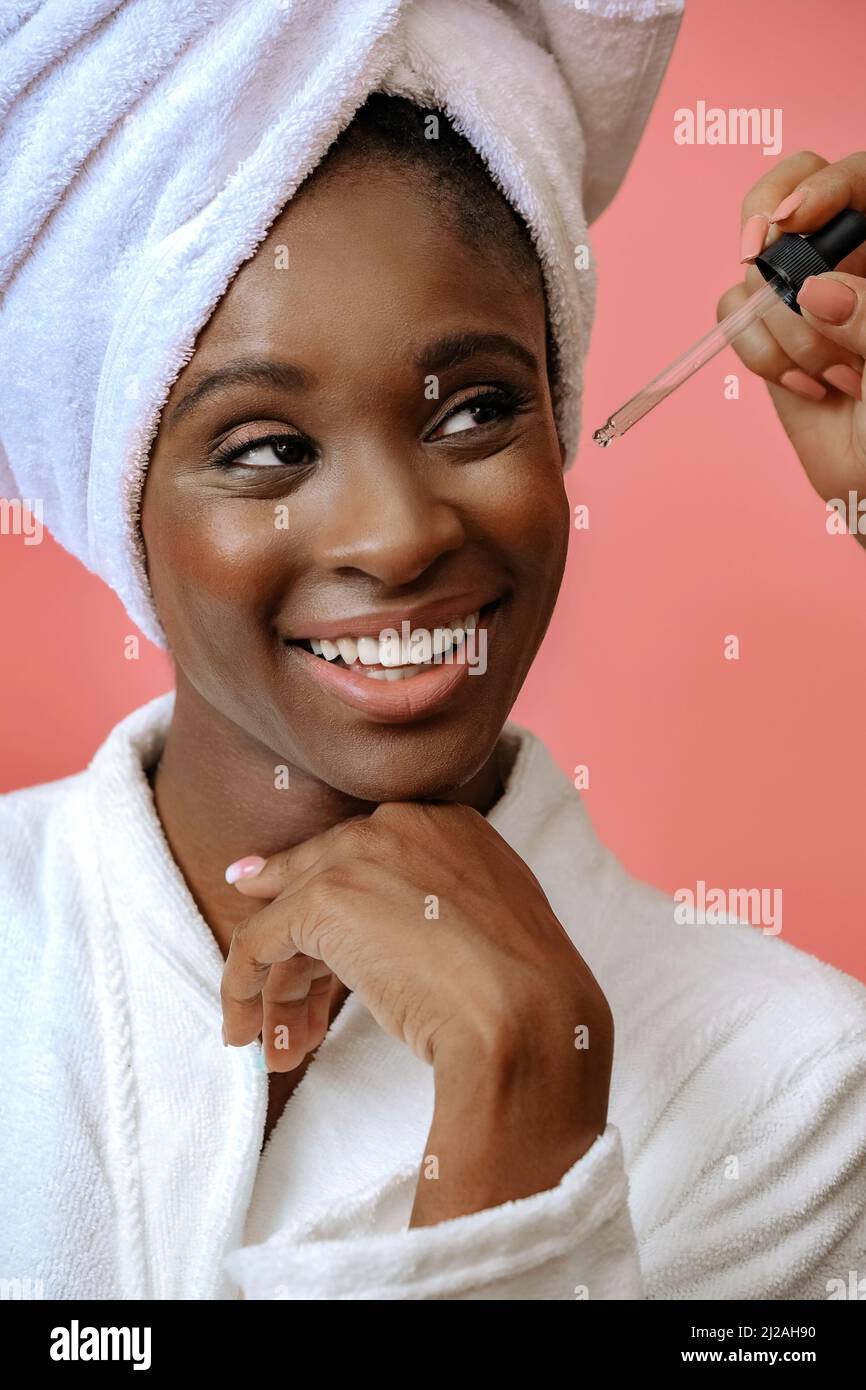 Ritratto di una giovane donna sorridente in accappatoio con asciugamano sulla testa che riceve il trattamento con pipetta su spazio rosa copia Foto Stock