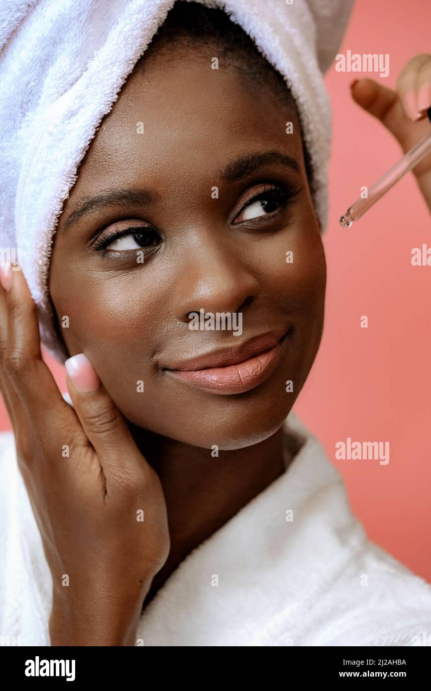 Primo piano ritratto di una giovane donna che pensa in accappatoio con asciugamano spa sulla testa che riceve il trattamento con pipetta su spazio rosa copia Foto Stock