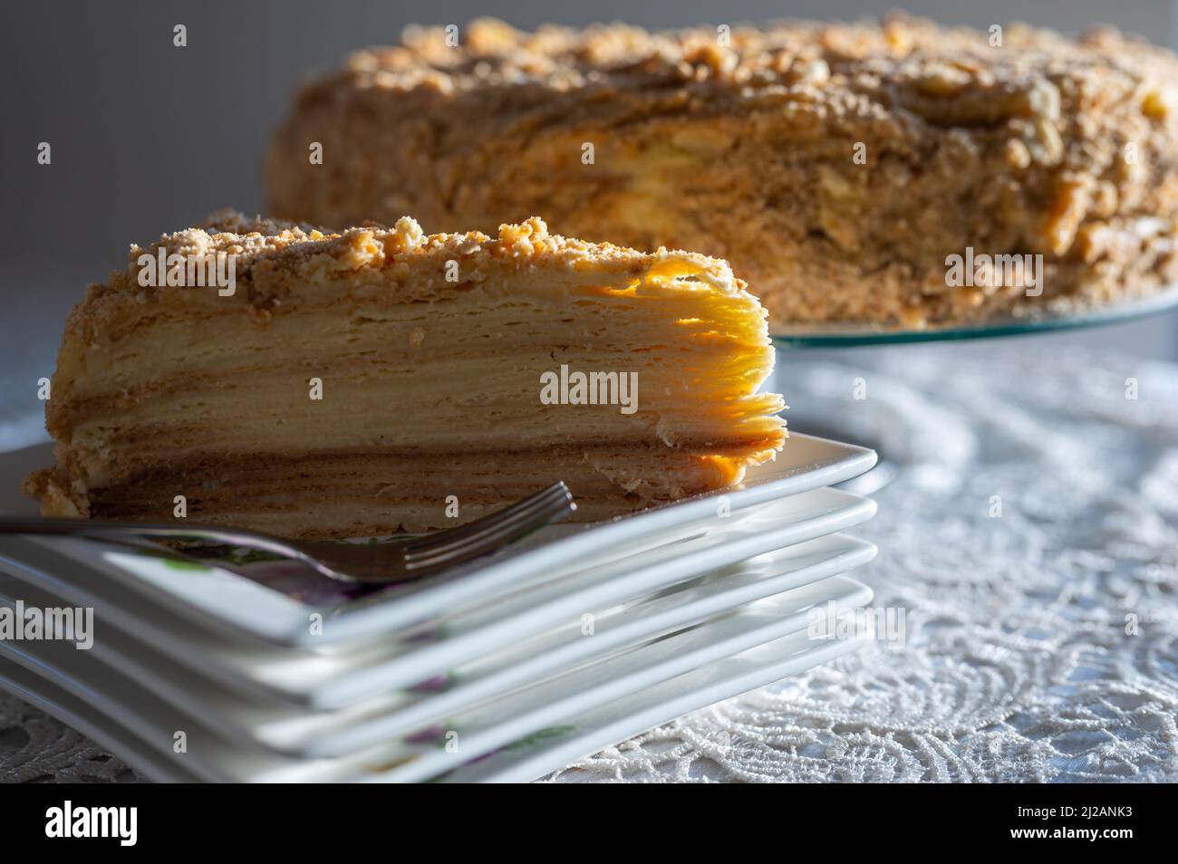 Prima fetta di famosa torta Napoleone servita su un piatto quadrato. Fetta triangolare di torta decorata con croccanti briciole illuminate dalla luce naturale del sole Foto Stock