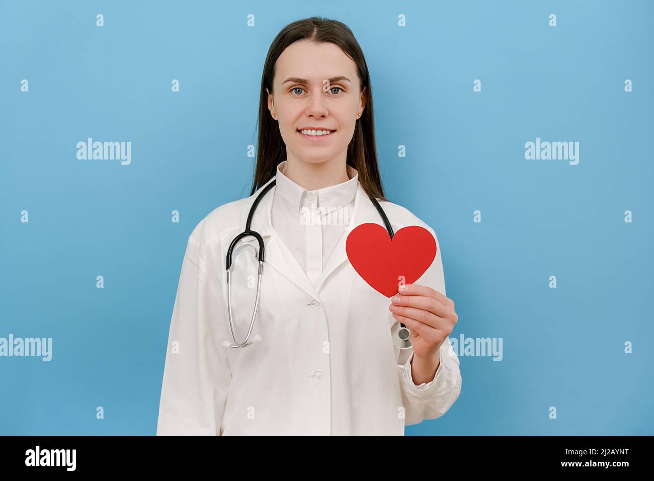 Ritratto di amichevole sorridente giovane medico in cappotto medico bianco professionale con stetoscopio che tiene rosso il cuore, guardando la macchina fotografica, posando l'isola Foto Stock