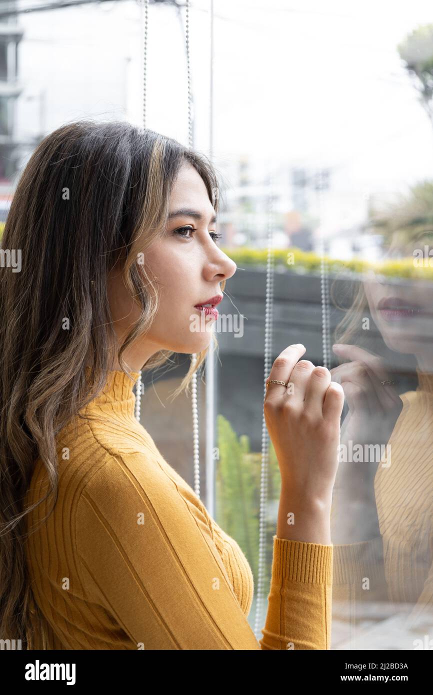 attraente donna latina con lunghi capelli biondi che guardano fuori la finestra della città, indossa trucco e vestiti colorati, bellezza e model lifestyle Foto Stock