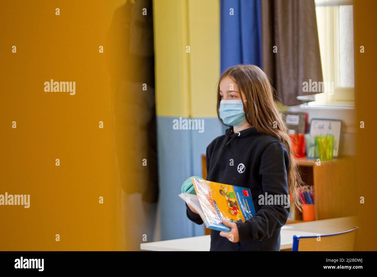 Scuola elementare durante il Covid-19, Coronavirus focolaio: Scolarella che indossa una maschera facciale che porta un libro in una classe Foto Stock