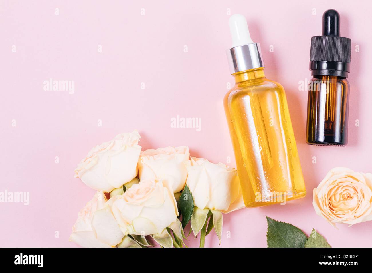 Prodotti cosmetici a base di estratto di rosa, siero e olio su sfondo rosa accanto a fiori freschi di rosa, piatto Foto Stock