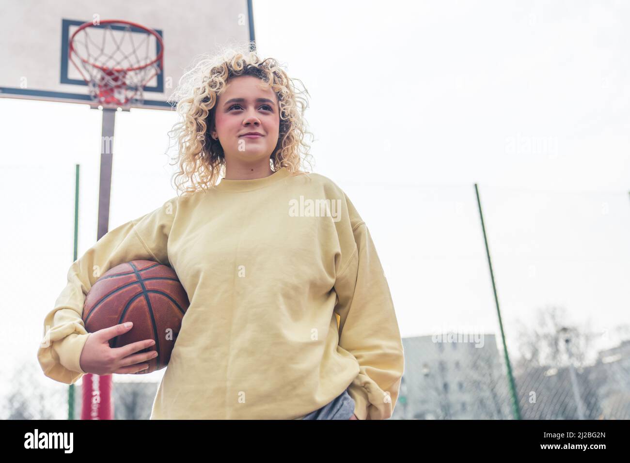 Bella giovane bionda capelli ricci donna mantenendo una palla di basket sul campo, cestino sullo sfondo, sorridente e guardando via, medio shot copia spazio . Foto di alta qualità Foto Stock
