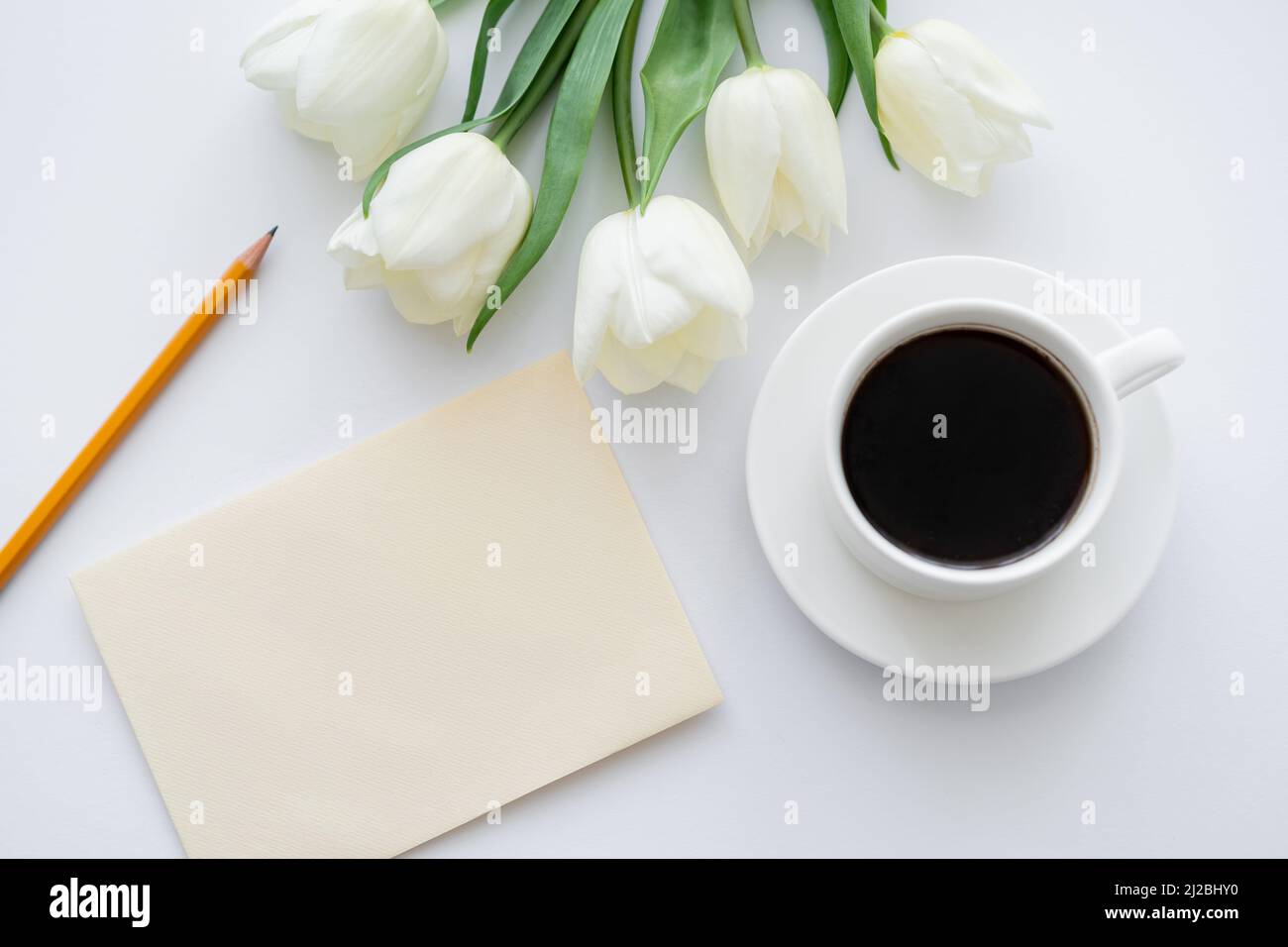 vista dall'alto della busta con matita vicino alla tazza con caffè e tulipani su bianco Foto Stock