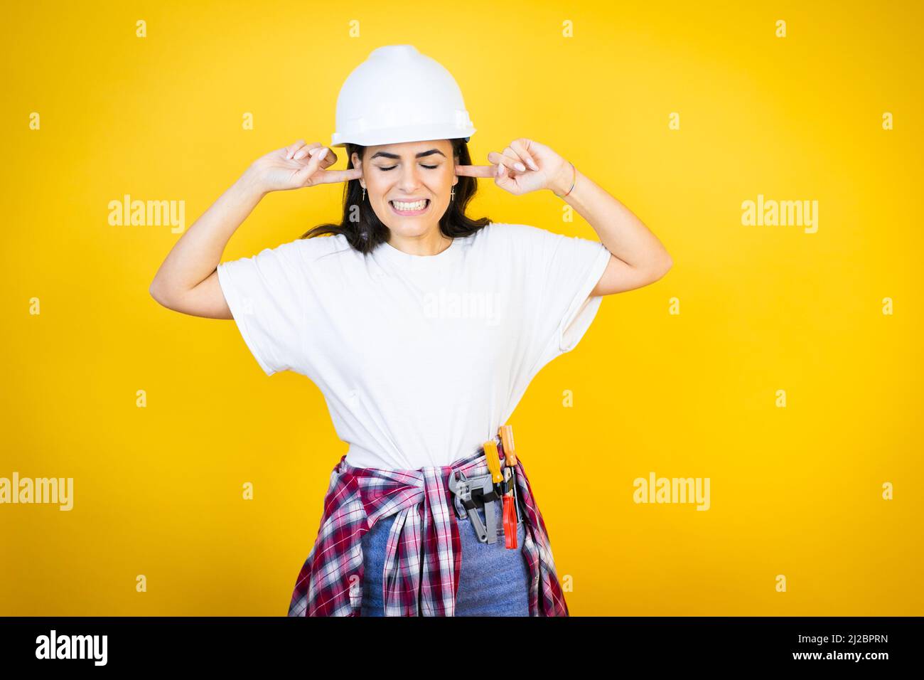 Giovane donna caucasica che indossa abiti hardhat e builder su fondo giallo isolato che copre le orecchie con le dita con espressione infastidita per il n Foto Stock