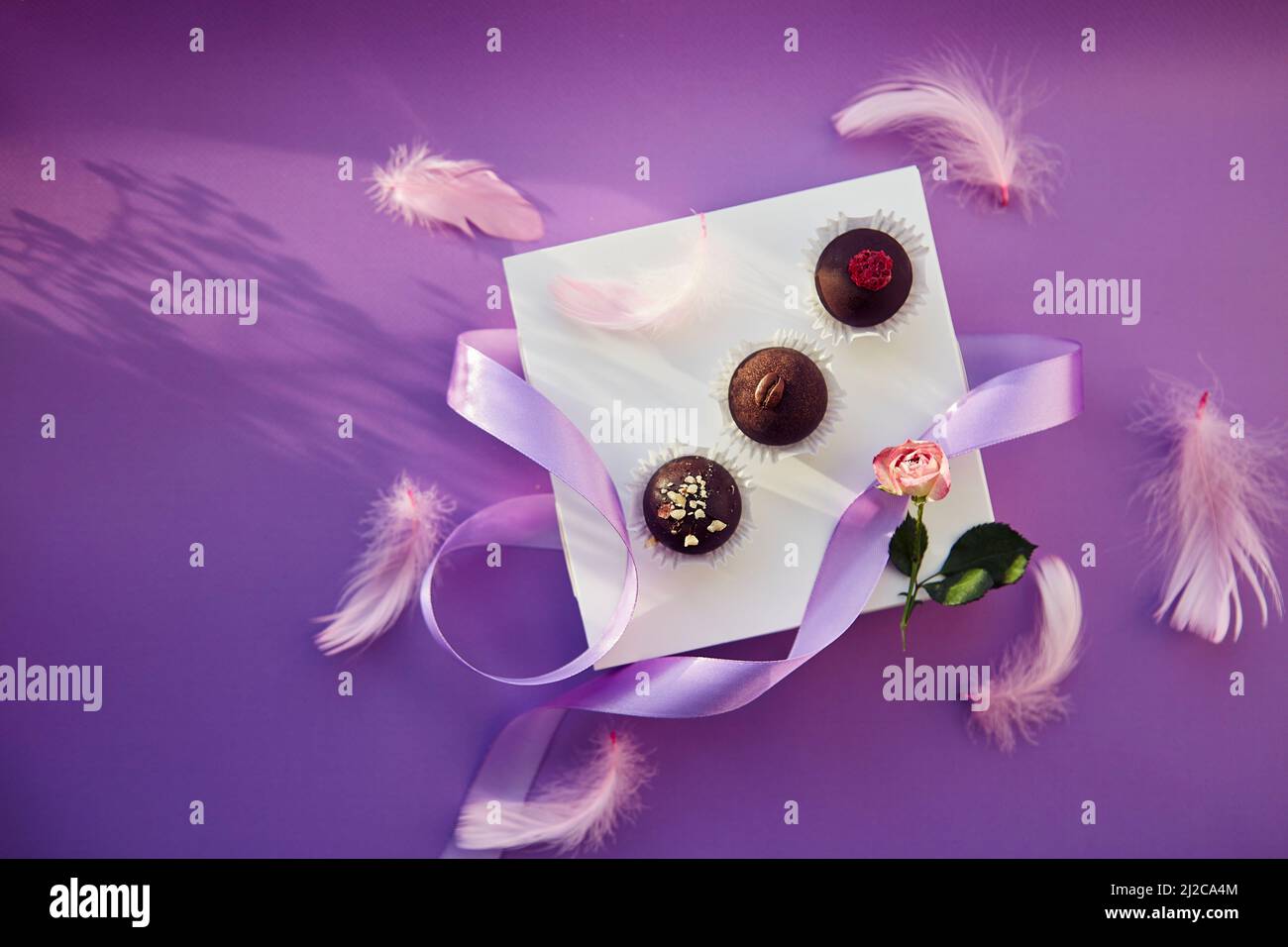 Caramelle al cioccolato festive. Delicate piume rosa con nastro viola. Sfondo con motivo di colore viola con ombre. Foto Stock