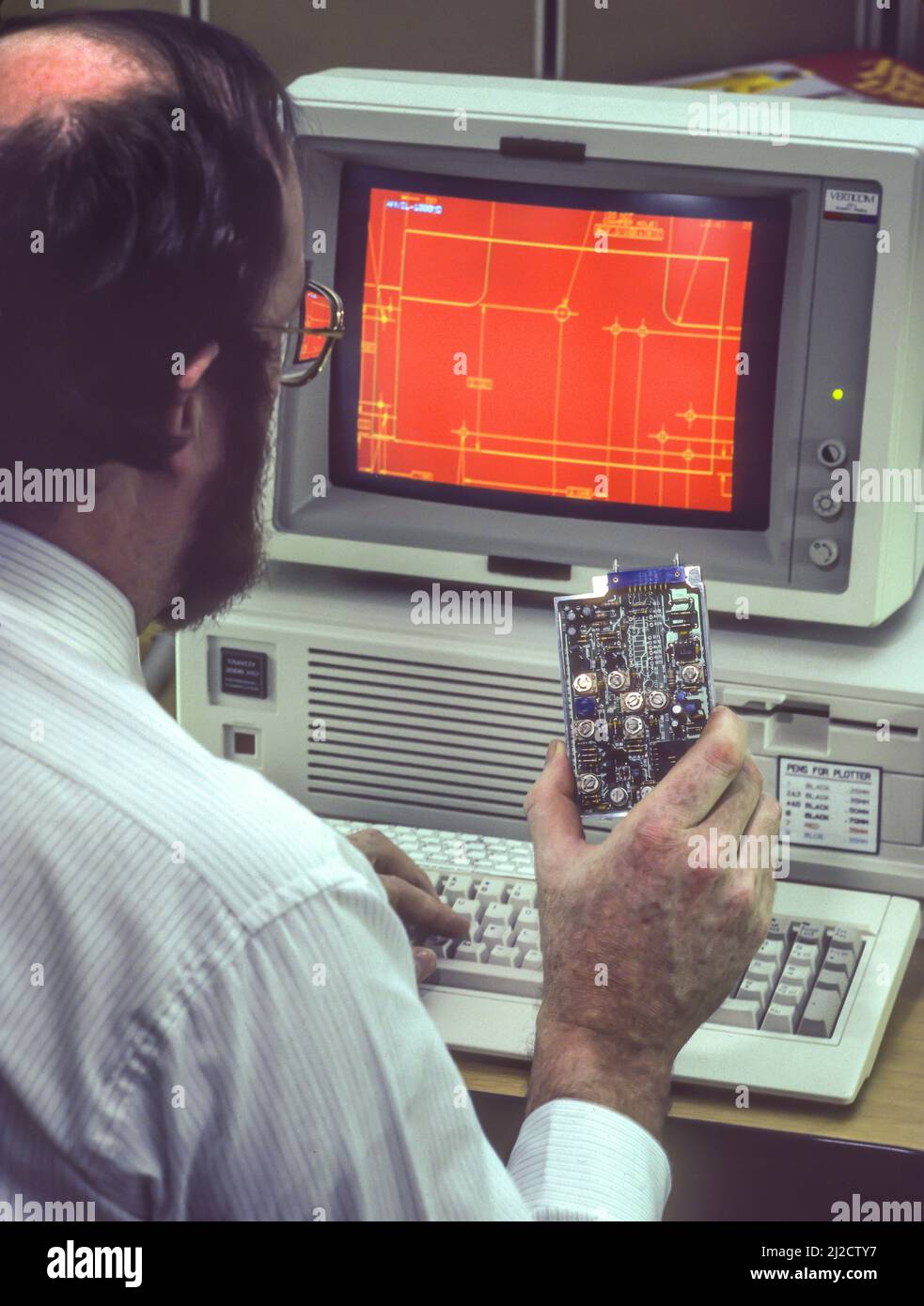 SYRACUSE, NEW YORK, USA - Man at computer lavora nel design computerizzato, febbraio 1987. Lo schermo del computer mostra lo schema della scheda tenuta in mano. Foto Stock