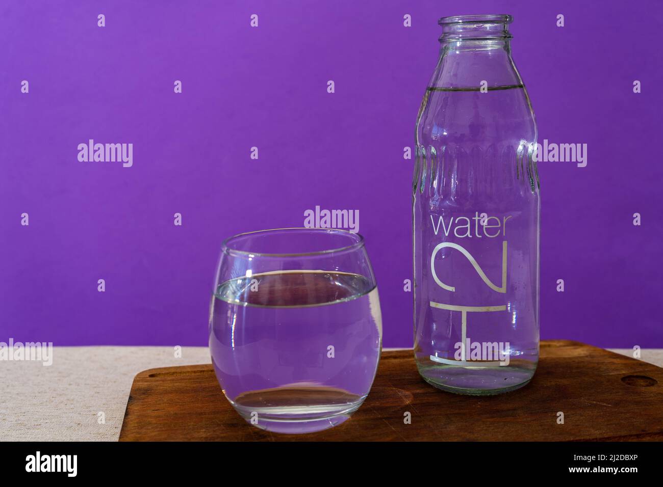 Bicchiere di vetro e bottiglia riempiti con acqua purificata. Sfondo viola. Foto Stock