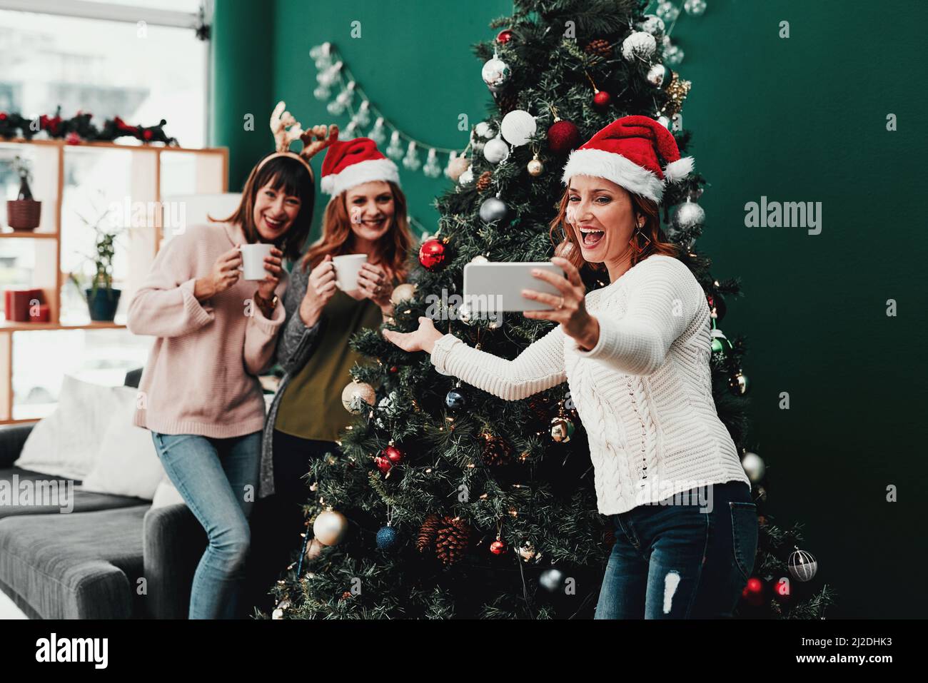 Stare più vicino all'albero. Scatto corto di tre donne attraenti di mezza età che prendono i autoritratti insieme ad un cellulare a casa durante il Natale Foto Stock