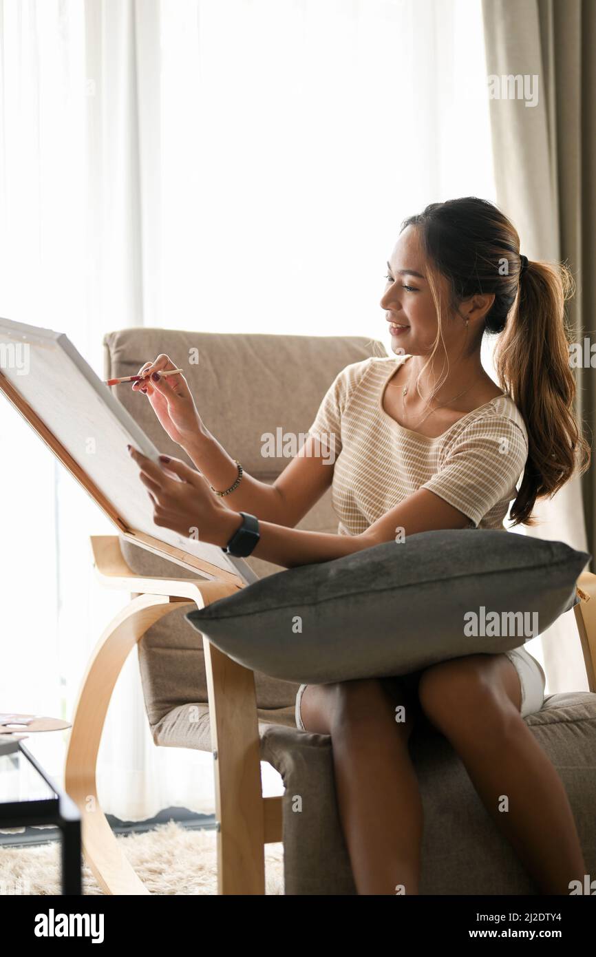 Elegante giovane donna asiatica seduta su una comoda poltrona e ammirando la sua bella opera d'arte su una tela Foto Stock