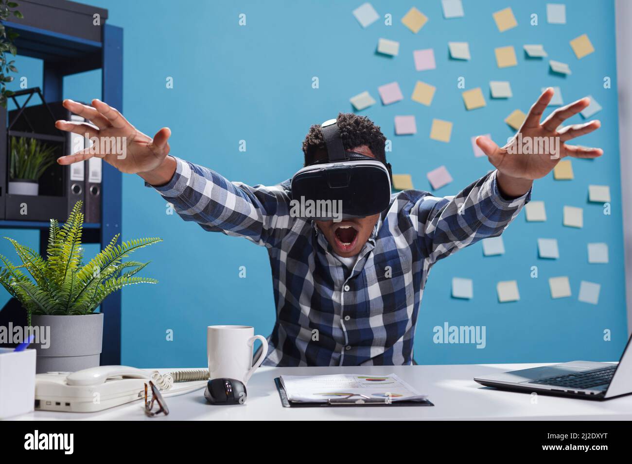 L'espressivo stupito impiegato dell'azienda afroamericana che gode della tecnologia di gioco di realtà virtuale. Uomo d'affari adulto che indossa un dispositivo di intrattenimento elettronico mentre si siede alla scrivania nello spazio di lavoro. Foto Stock
