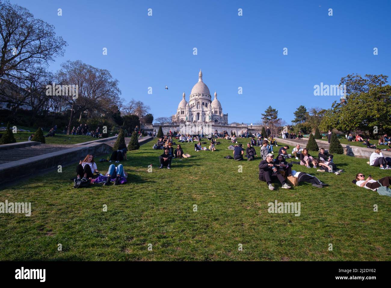 Parigi, Francia - 22 marzo 2022 : la Basilica del Sacro cuore di Parigi, comunemente conosciuta come Sacre - Coeur e una folla di persone che siedono sull'erba Foto Stock