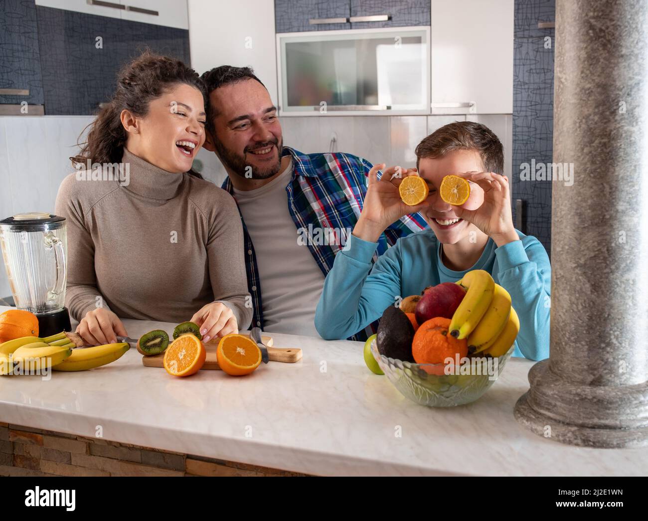 Madre e padre in piedi in cucina ridendo a loro figlio che sta ingannando intorno mentre tiene le fette arancioni sugli occhi Foto Stock